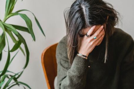 Avoiding Mistakes - Unrecognizable young upset female millennial with dark hair in stylish sweater covering face with hand while siting on chair at home