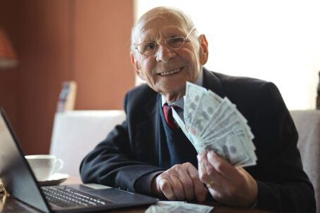 Smart Investing - Happy senior businessman holding money in hand while working on laptop at table