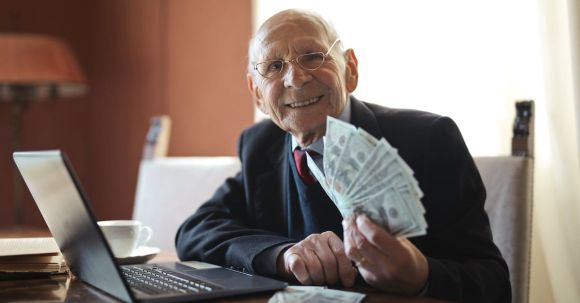 Smart Investing - Happy senior businessman holding money in hand while working on laptop at table